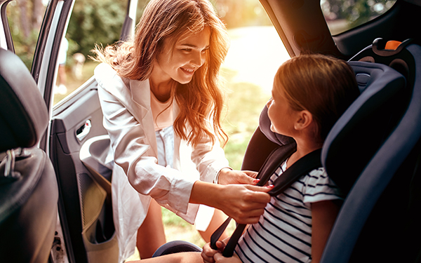 A divorced parent is smiling at her happy daughter, as she buckles her in a carseat after a coparenting transition
